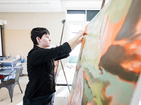 An art student leaning in to paint details on a large colorful canvas set on an easel.