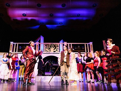 Actors in elaborate costumes performing a theatrical play on a brightly lit stage set.